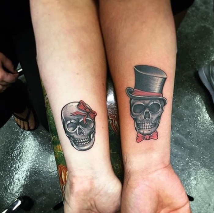 top hat in black and red, and a red bow tie, on a skull, tattooed near the wrist of an arm, his and hers tattoos, the arm next to it also features a skull tattoo, decorated with a red head bow
