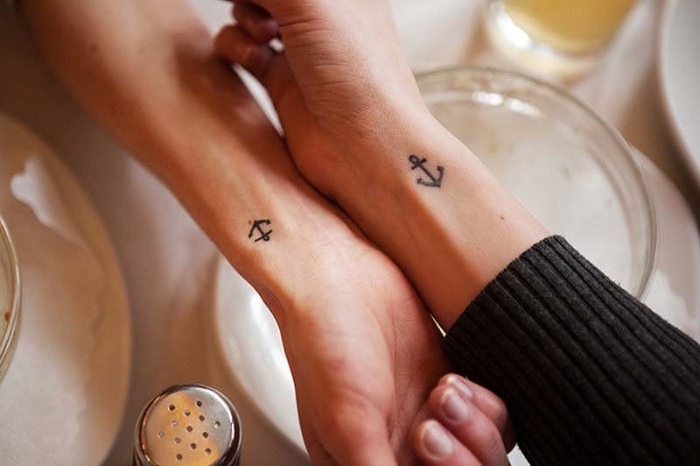 tiny black anchors, matching tattoos, done near the wrists of two hands, raised over a table