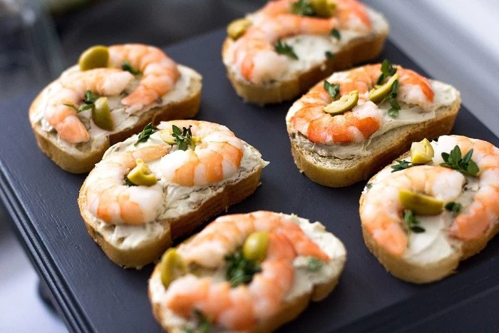six pieces of bread, with cream cheese spread, topped with prawns, green olives and parsley