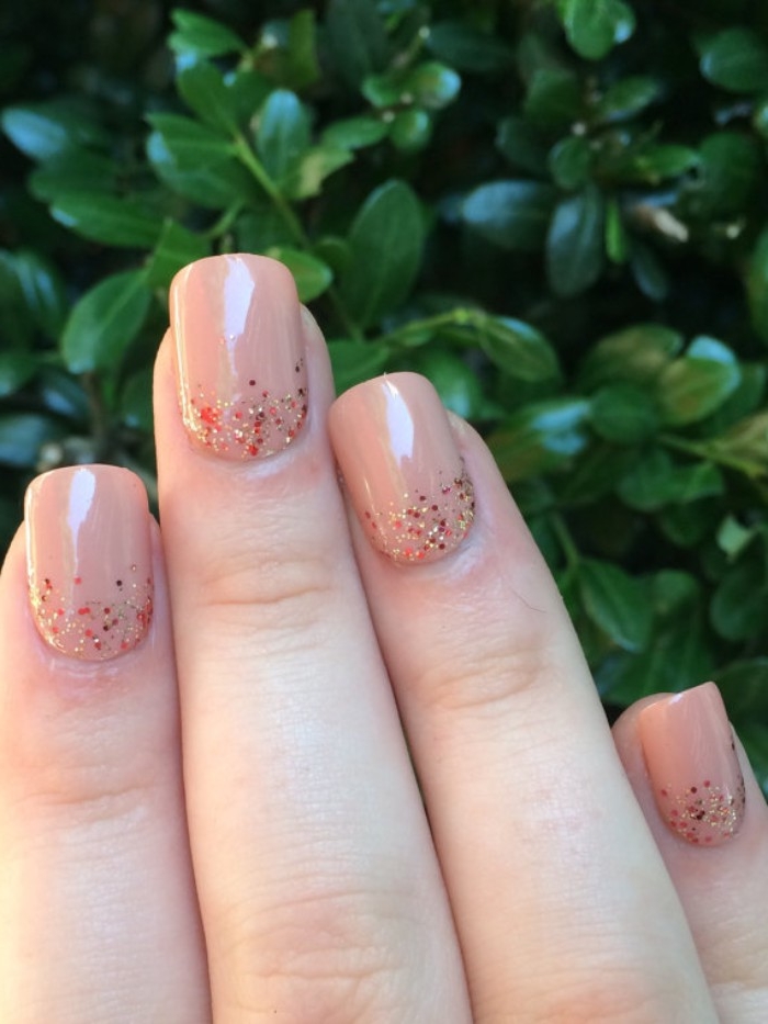 fine glitter in red and gold, decorating the base, of four square nails, painted in a nude nail polish, green tree in the background