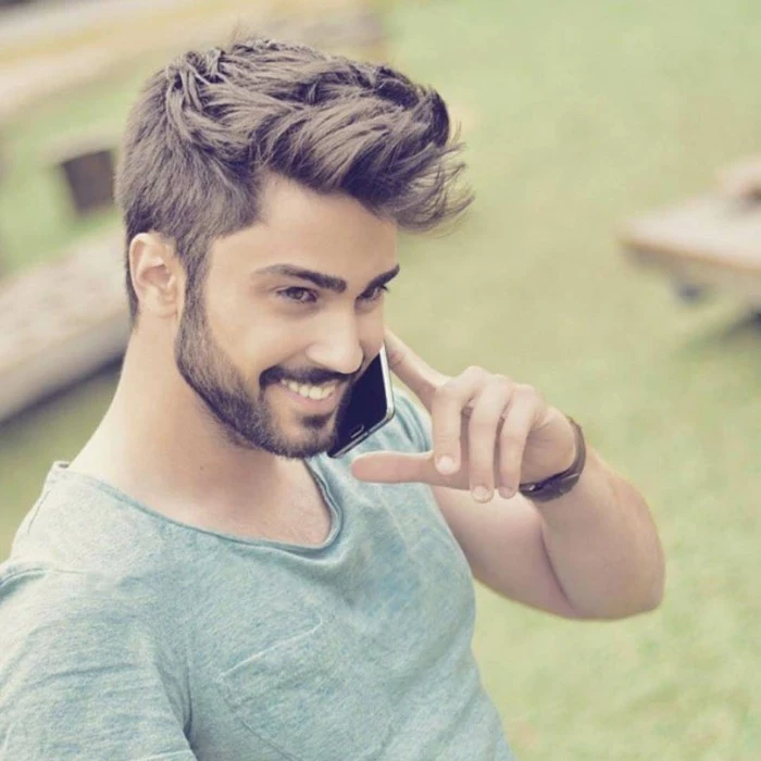 trendy haircuts for men, smiling young man, with a moustache and a short beard, holding a phone to his face, brunette textured cut