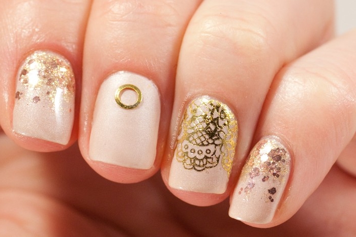 lace-like motifs in gold, gold glitter flakes, and a small ring-like sticker, on nude nail polish, decorating four square nails