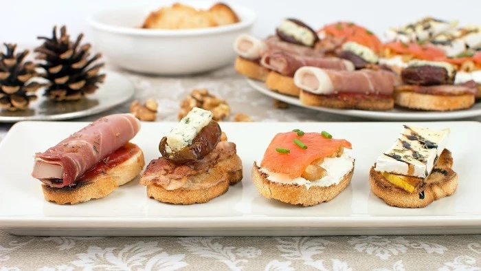 holiday horderves ideas, four slices of toasted bread, topped with cheese, smoked salmon and chives, prosciutto and other foods