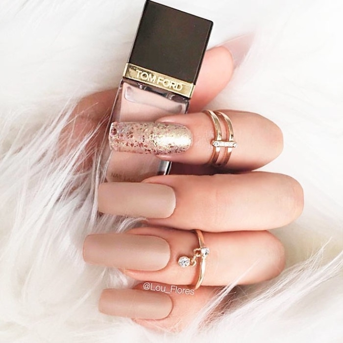 very long and square, nude matte nails, one decorated with metallic glitter, on a hand with two golden rings, holding a bottle of nail polish