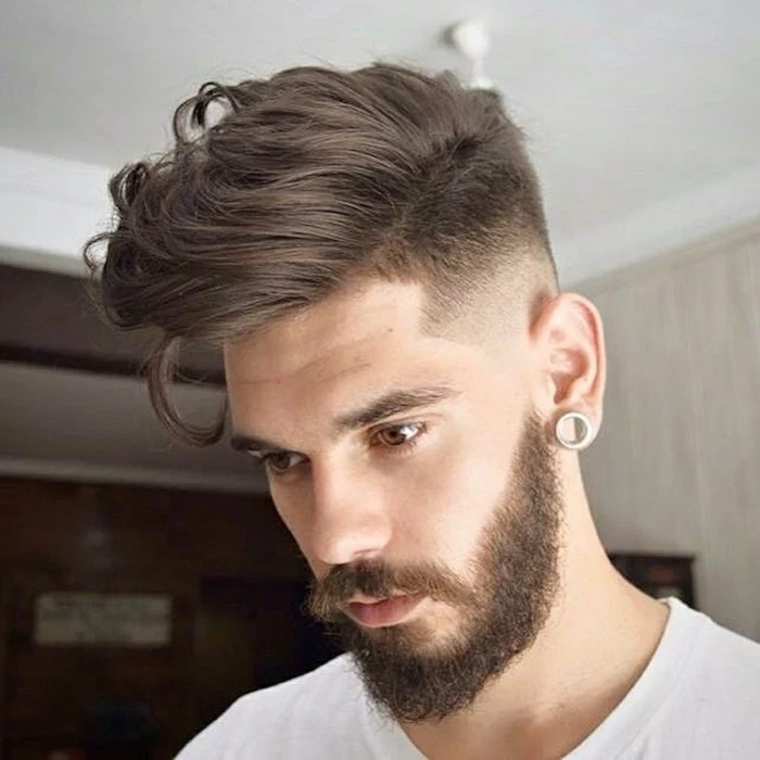 wavy chocolate brunette hair, styled to one side, with a connected undercut, modern haircuts for men, worn by a bearded young man, with a mustache and an earring