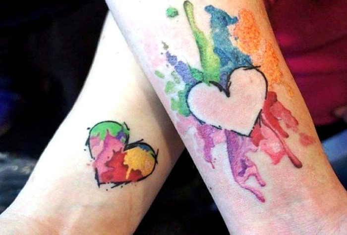 multicolored splashes with watercolor effect, and two heart shapes, matching tattoos for couples in love, done near the wrists of two arms