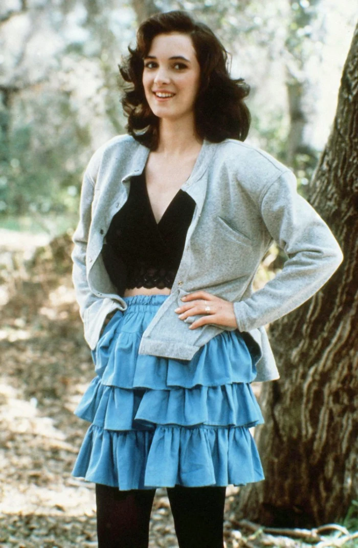 rara skirt in blue, worn with a black crop top, and a pale grey cardigan, by a young winnona ryder, 80's fashion pictures, smiling with hands on her hips, dress code nostalgia