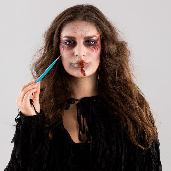 scary face paint, painting small thin veins, in blue and red, near the corners of the eyes, of a young brunette woman, wearing zombie makeup
