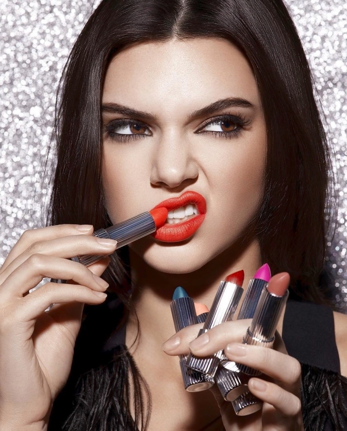 scowling young woman, with dark brunette hair, holiday makeup 2017, applying red lipstick with one hand, while holding five lipsticks, in different colors, with the other hand 