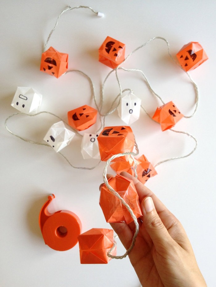 using sticky tape, to attach white and orange, paper origami shapes, with black hand-drawn jack-o-lantern faces, on a string of decorative lights