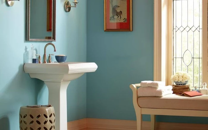 robin's egg blue walls, in a bright room, with a white sink, and a settee in pale beige and white, small bathroom paint colors, framed artwork and a window