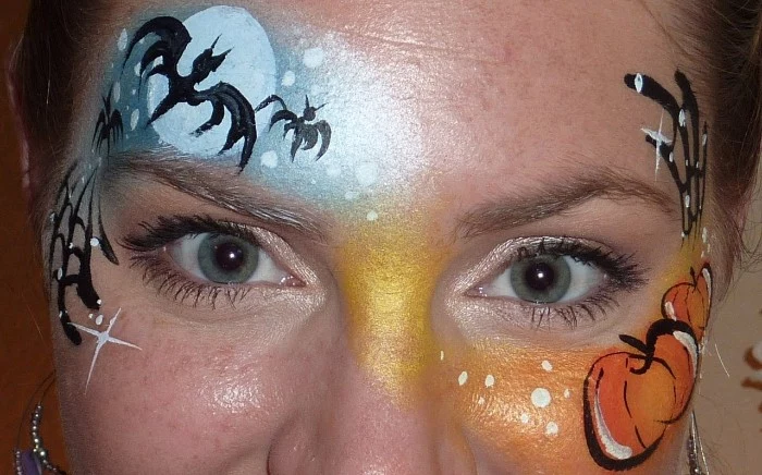pumpkins and bats, painted on the forehead of a woman, with blue eyes and freckles, halloween face paint ideas for adults, pale blue and orange paint