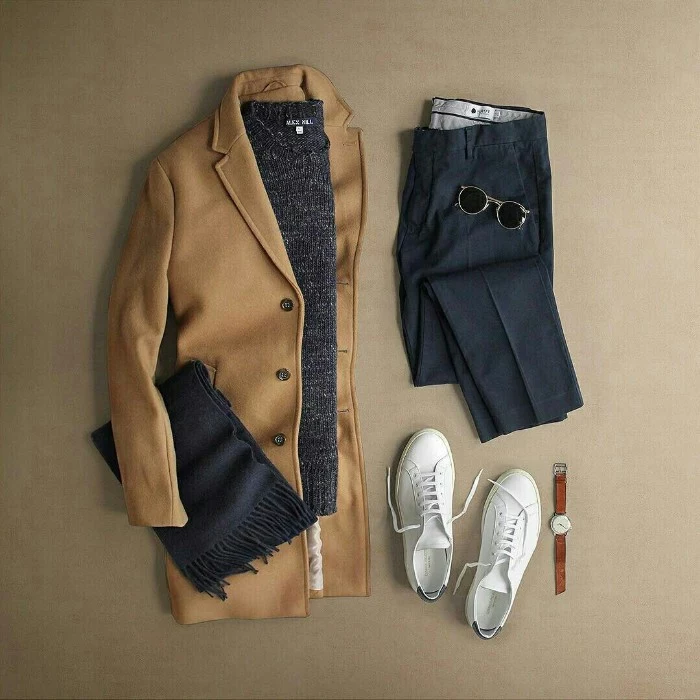 camel colored woolen coat, dark grey sweater, a folded pair of black trousers, white sneakers and a watch, dark grey woolen scarf, and a pair of black sunglasses, wardrobe essentials