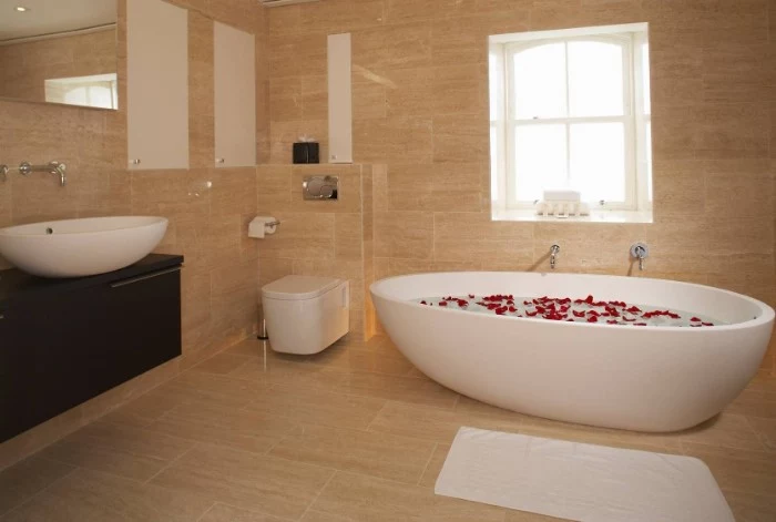rose petals floating, in an oval white bathtub, filled with water, inside a room, covered with beige tiles