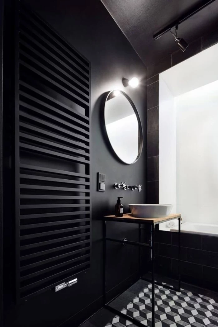 lightbulb glowing over a round mirror, mounted on a black wall, near a tall black towel rack, white grey and black tiled floor, white bath area