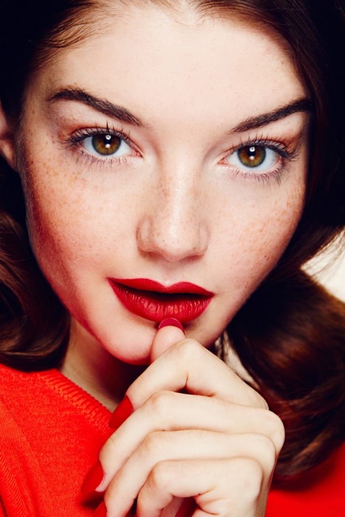 freckled brown-eyed young woman, with brunette hair, wearing a bright red top, red nail polish, and matching red lipstick