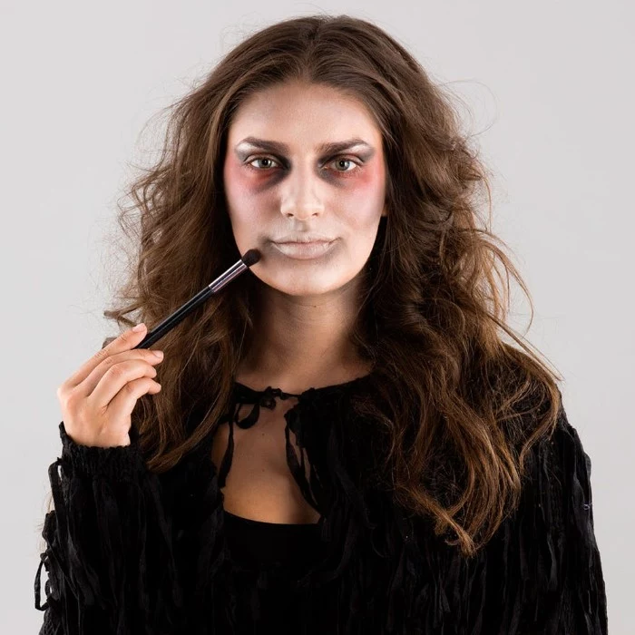 outlining the mouth of a young, brunette woman, using grey makeup and a brush, zombie face paint, and a black tasseled costume