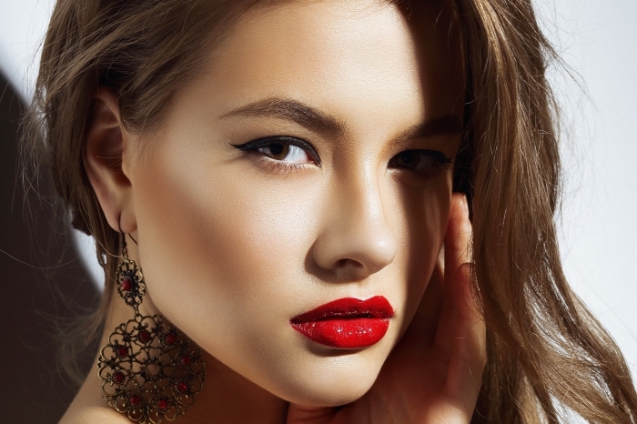 glossy red lipstick, and subtle black eyeliner, christmas makeup ideas, worn by a young brunette woman, with large ornamental earrings, and subtle blush