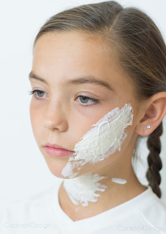 girl with blue eyes, and brunette hair, braided in pigtails, wearing a white t-shirt, special effects makeup wax on her cheek and neck, how to create scary face paint