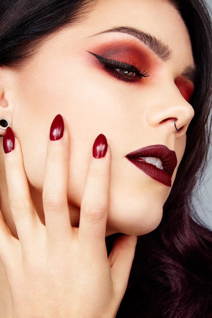 eyeliner in black, and red eye shadow, worn by a pale young woman, with burgundy red lipstick, and a small nose ring, christmas eye makeup, almond shaped nails, painted in dark red nail polish