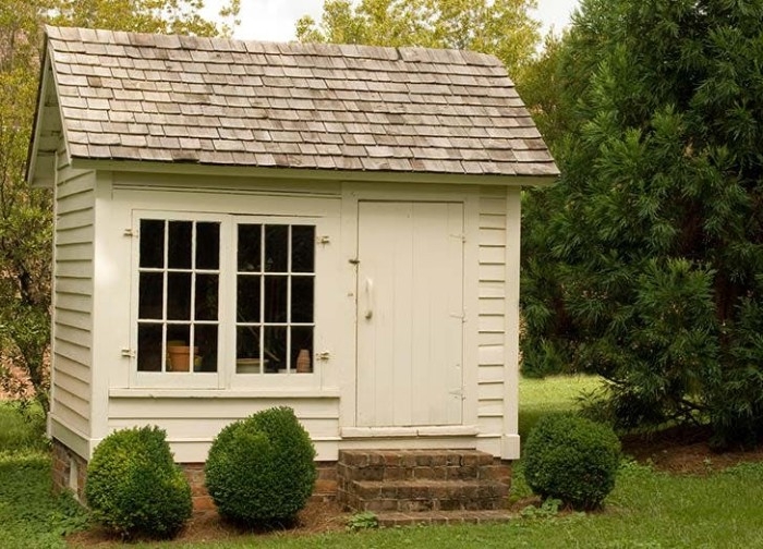 simple small white shed, with grey tiled roof, a double window, and a white door, she shed images, three green shrubs, grass and trees surrounding it 