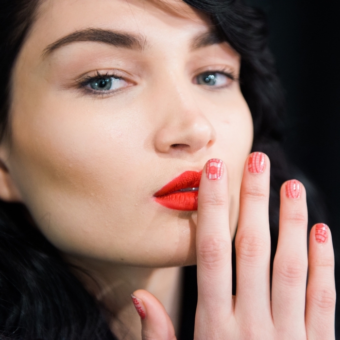 orange lipstick worn by a pale young woman, with black wavy hair, and blue eyes, short manicure with red and white nail polish