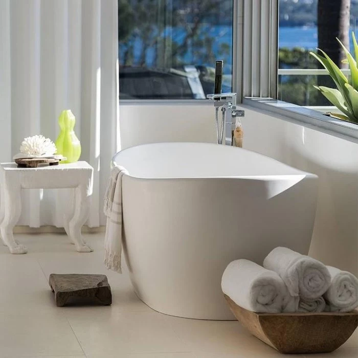 oval white bathtub, inside a room with two windows, and white curtains, master bathroom ideas, a wooden dish with rolled up towels, a small chair with toiletries