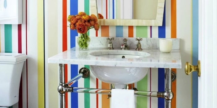 multicolored stripes on a wall, behind a marble sink, decorated with a candle, and a vase with orange flowers, best bathroom paint colors, mirror in an ivory-colored frame