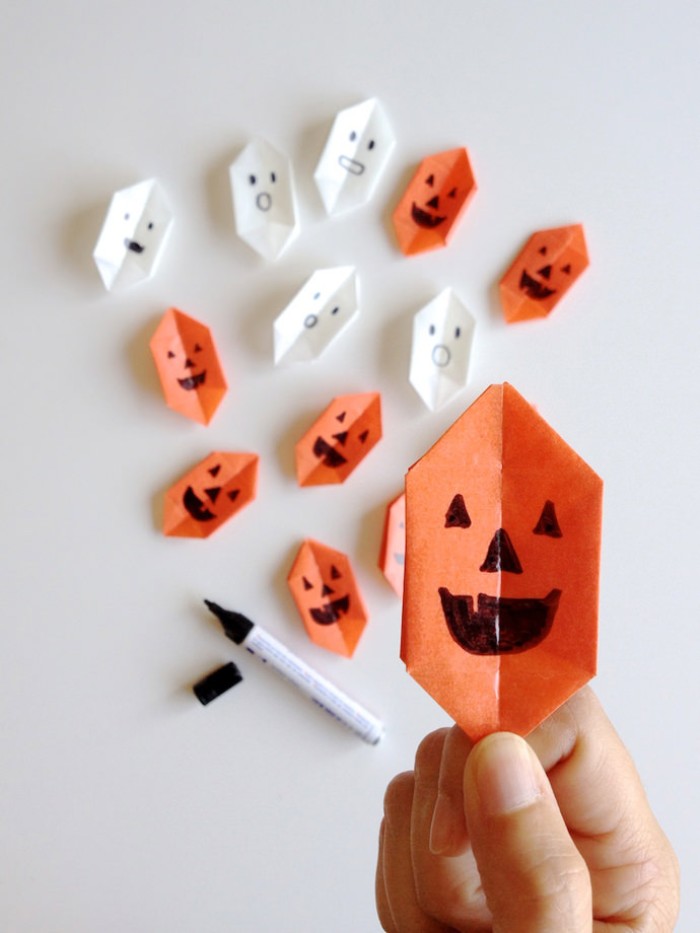 smiling jack-o-lantern face, drawn with black permanent marker, on an orange, paper origami shape, more white and orange shapes, and a marker, in tne background