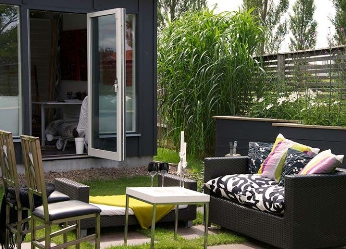 coffee table in white, dark grey rattan couch, and a few chairs, in front of a modern she shed, with an open door