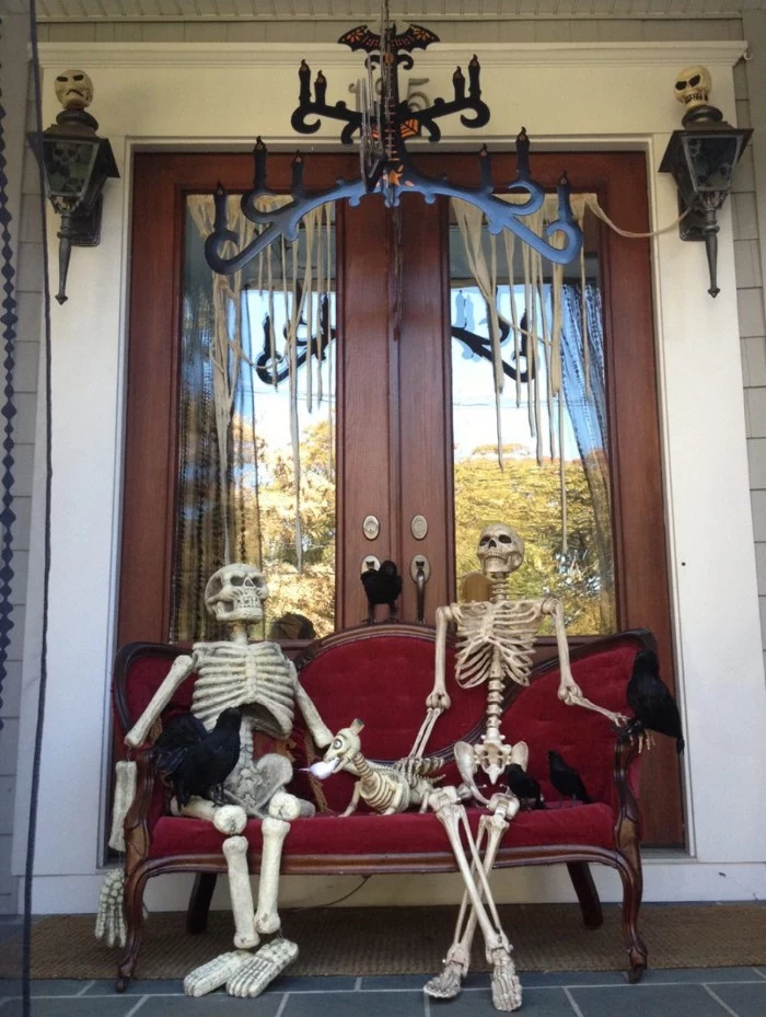 two fake skelettons, sitting on an antique, red velvet settee, with several fake crows, and a skeleton dog, scary outdoor halloween decorations, in front of a double door, decorated with two skulls, and fake cobwebs