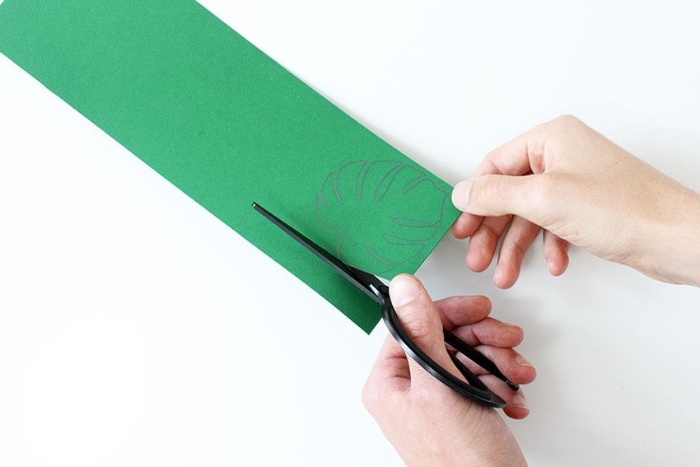 scissors held by a pale hand, cutting along the lines of a palm leaf shape, drawn in pencil, on a piece of green card, held by another hand, how to decorate a bedroom, using arts and crafts