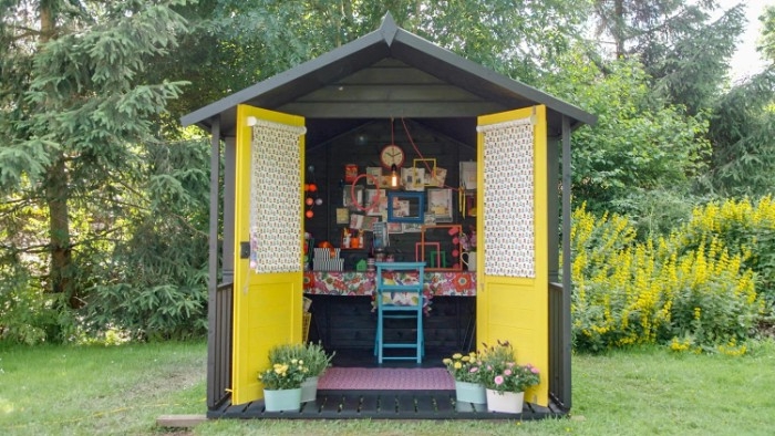 wide open yellow doors, attached to a small, dark grey she shed, desk and chair, and lots of colorful frames visible inside