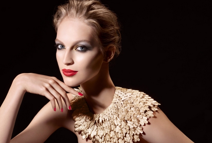 collar-like decoration made of gold, worn by a slim young woman, with short slicked back blonde hair, and holiday makeup, red lipstick and pale blue eye shadow 