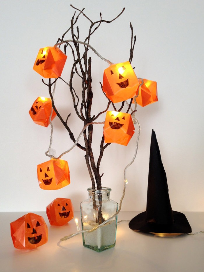 lit pumpkin fairy lights, string lights covered in orange, paper origami shapes, with hand-painted jack-o-lantern faces, placed on dried tree branches in a vase 