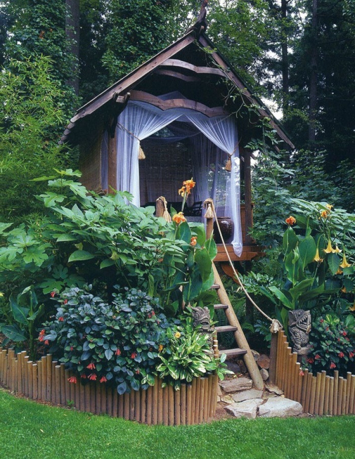 sheer curtains in white, on a small tree-house style shed, accessible via a wooden ladder, and surrounded by various shrubs and flowers