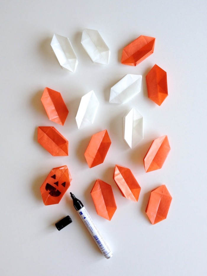 fifteen paper origami shapes, in orange and white, one shape has a hand-drawn, jack-o-lantern face, done in black marker