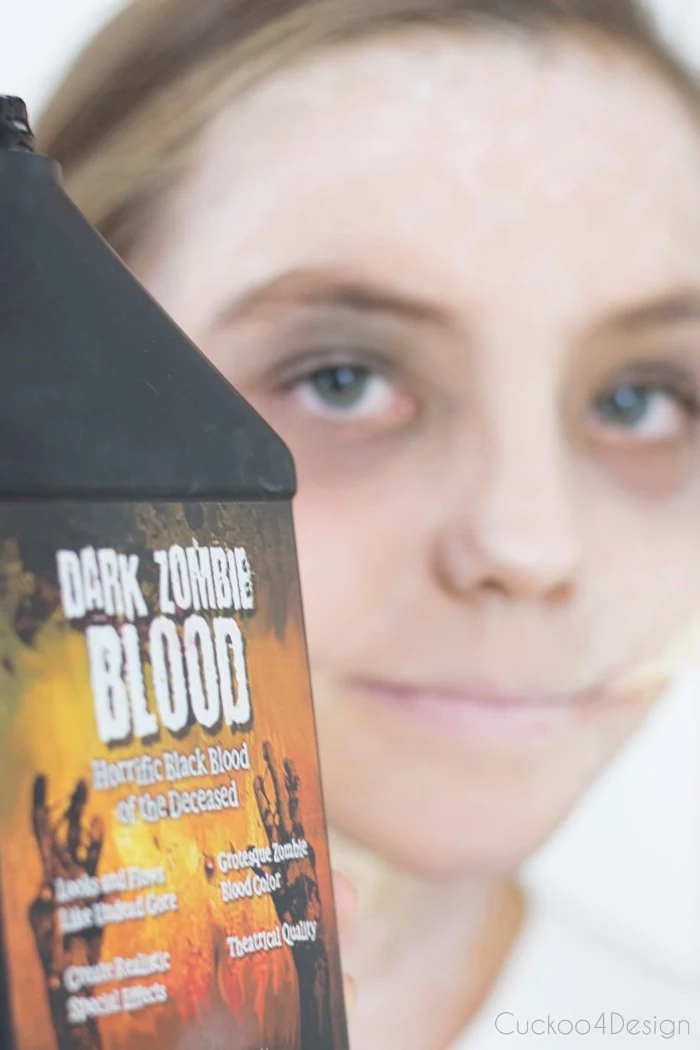 facepaint ideas, close up of a black bottle, with the inscription dark zombie blood on the label, smiling young girl, with pale makeup in the background