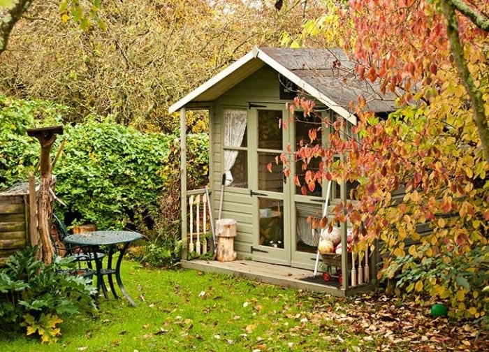 autumn leaves on multiple trees, surrounding a small pastel green hut, she shed ideas, glass double doors, and small narrow windows