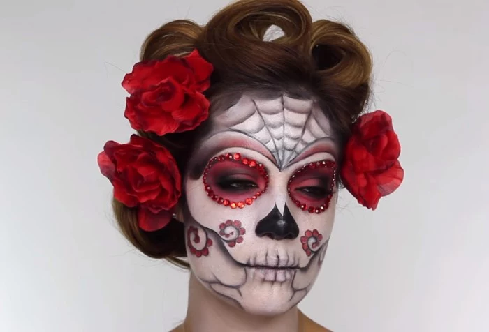 finished sugar skull face paint, worn by a young woman, with curled brunette hair, decorated with three red faux flowers, halloween face paint, in white and black and red, decorated with rhinestones