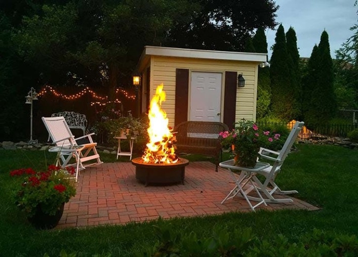 light yellow small hut, in a garden with flowers and trees,shed ideas, two white garden chairs, a small table with a potted plant, and a firepit