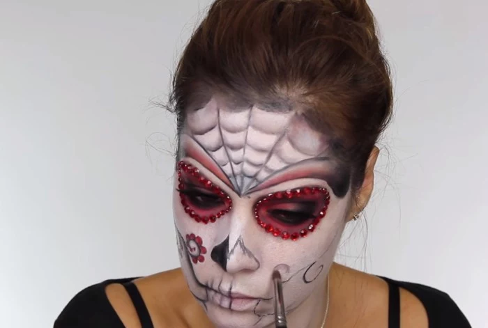 drawing a floral motif, on the face of a young woman, covered in white, and pink paint, with black details, skeleton face paint, creating a sugar skull costume