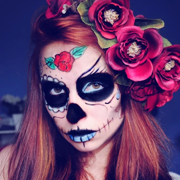 several fake red flowers, decorating the red hair, of a young woman, wearing a sugar skull face paint, in black and blue, red and green