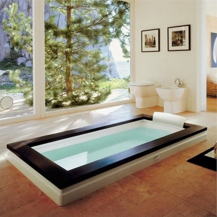 fir tree visible from the large windows, of a bright room, containing a large rectangular batthub, in black and white, spa like bathrooms, beige tiled floor, two framed photos