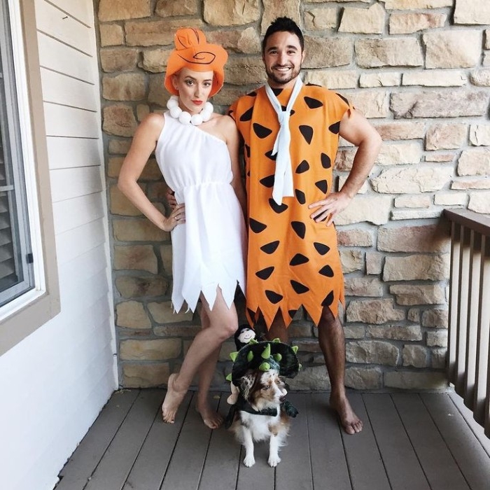 orange tunic with black pattern, and a white tie, worn by a smiling man, hugging a woman, in a white off-the-shoulder dress, with an orange paper wig, small dog dressed as a dinosaur, duo halloween costumes, inspired by the flintstones