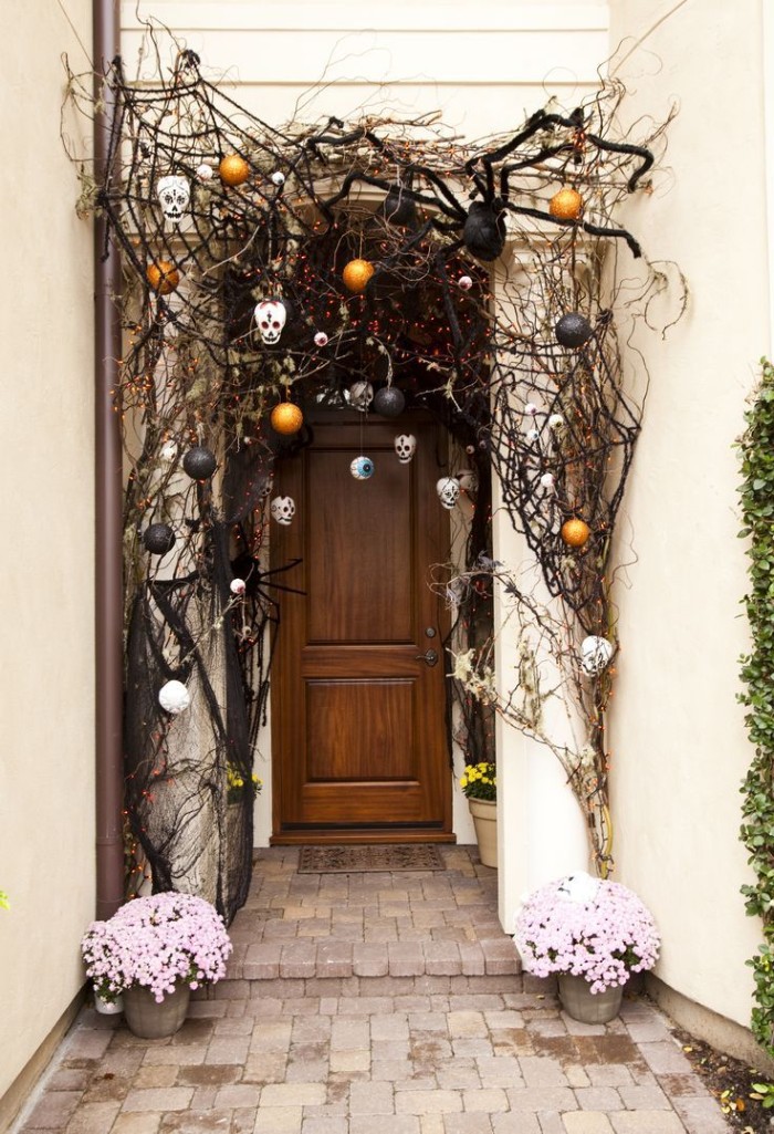 giant black spider ornament, black cobwebs and small white skulls, scary halloween decorations, near a front door, decorated with dried twigs, and black and orange baubles