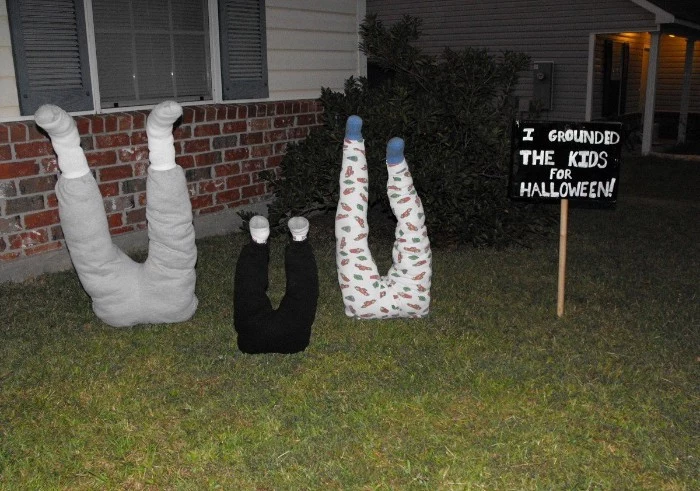 three sets of legs, in different sizes, dressed in trousers and socks, sticking out from the ground in a garden, scary outdoor halloween decorations, i grounded the kids for halloween, written in white, on a black sign