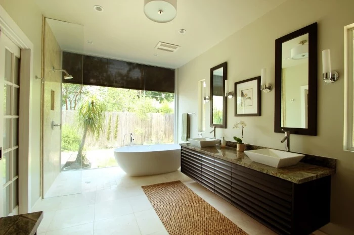 long dark brown cubpoard, with a grey marble counter top, containing two white sinks, master bathroom remodel, white bathtub near a large window, overlooking a garden