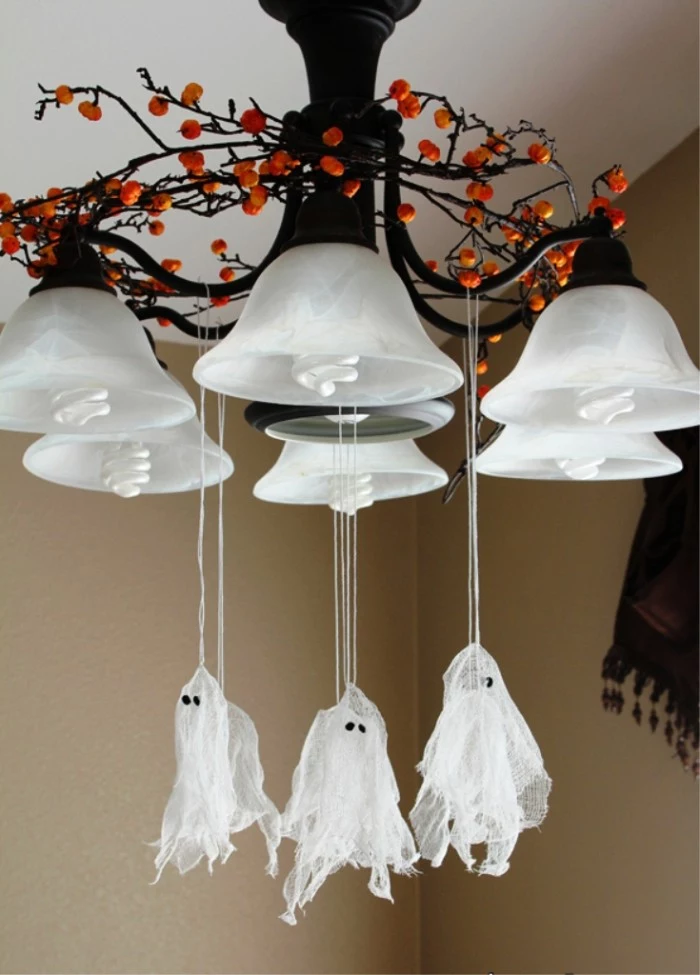 several small ghost ornaments, made from white gauze, scary halloween decorations, hanging on pieces of white string, from a chandelier, decorated with a wreath, made from brown twigs, with orange berries