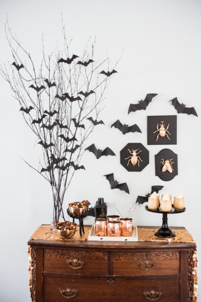 antique wooden cupboard, decorated with small dishes and candles, in black and gold, and a clear vase, containing dried branches, with small black bat shapes, insects ornaments and paper bats on the wall nearby